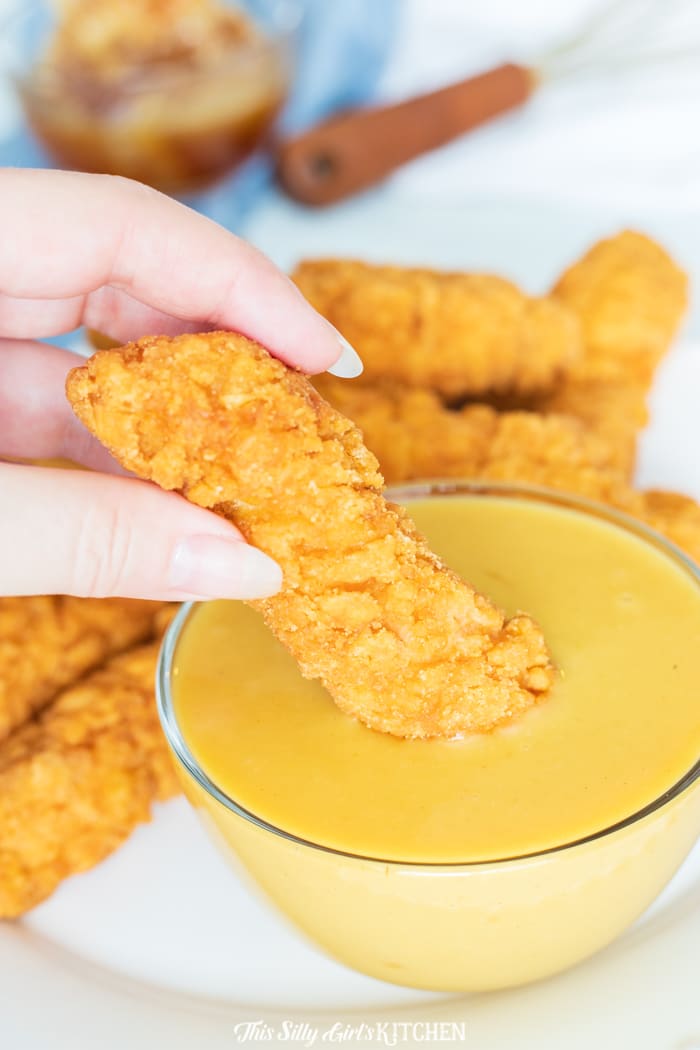 A crispy chicken tender being dipped into a bowl of Honey Mustard sauce.