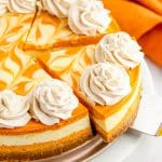 A whole swirled pumpkin cheesecake topped with piped cinnamon whipped cream rosettes on a white cake stand.