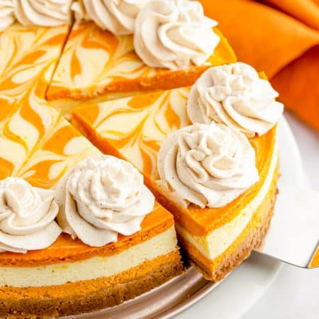 A whole swirled pumpkin cheesecake topped with piped cinnamon whipped cream rosettes on a white cake stand.