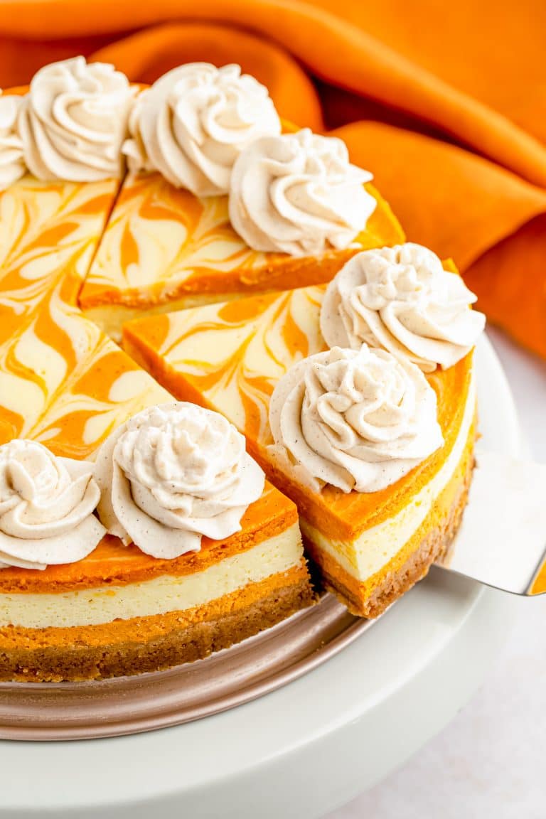 A whole swirled pumpkin cheesecake topped with piped cinnamon whipped cream rosettes on a white cake stand.