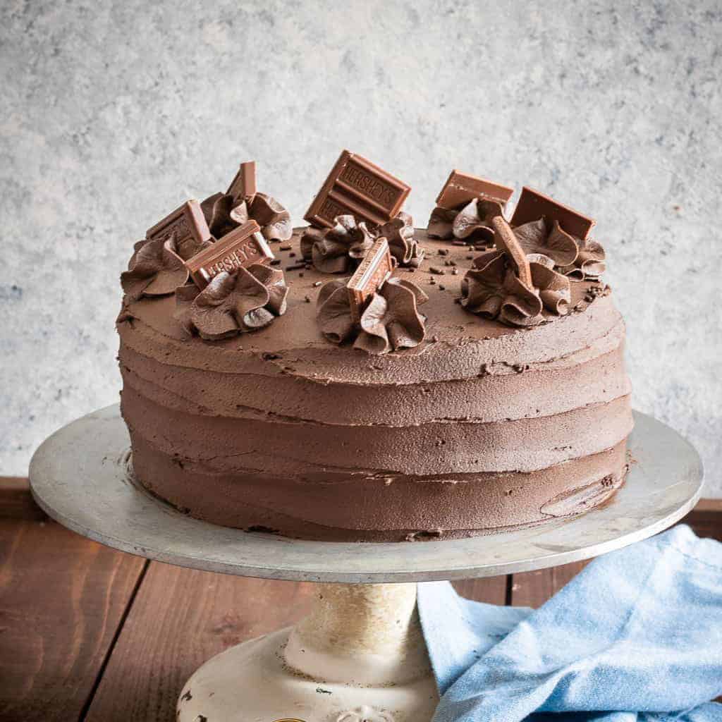 A whole frosted chocolate layer cake topped with chocolate shavings on a cake stand.