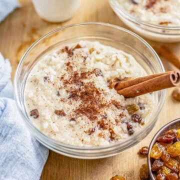 Bowl of Rice Pudding with Cooked Rice topped with cinnamon and a cinnamon stick with raisins on the side.