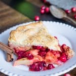 A slice of Apple Cranberry Pie on a white plate with cinnamon sticks and fresh cranberries around it.