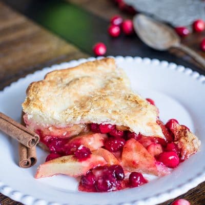 A slice of Apple Cranberry Pie on a white plate with cinnamon sticks and fresh cranberries around it.