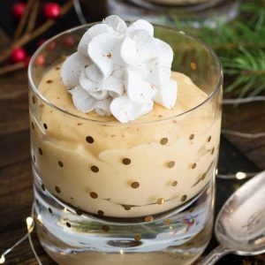 A glass of homemade Eggnog Pudding topped with cinnamon whipped cream on a holiday table.