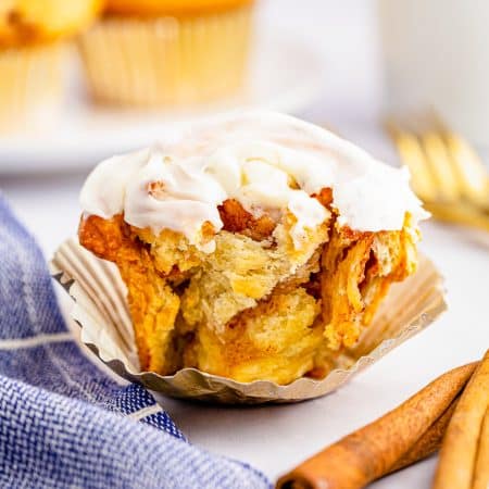 A frosted homemade Cinnamon Roll in a foil liner on a blue linen with cinnamon sticks.