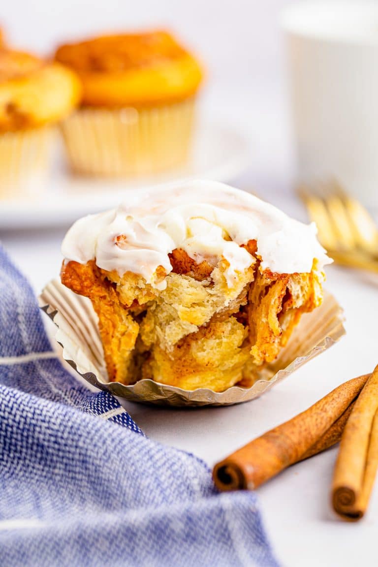 A frosted homemade Cinnamon Roll in a foil liner on a blue linen with cinnamon sticks.