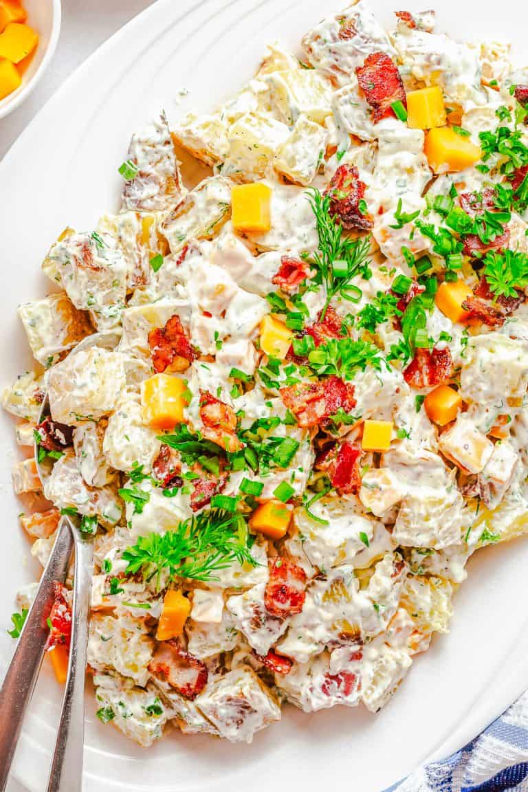 A platter of Ranch Potato Salad topped with crispy bacon, cheddar cubes, fresh dill, and scallions.