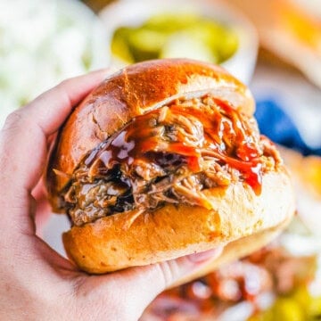 A hand holding a saucy slow cooker BBQ beef sandwich on a brioche bun.
