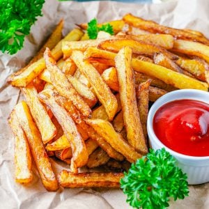 Square image of French Fries on brown parchment with parsley and ketchup.