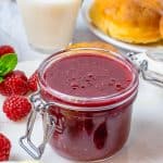 A glass jar of glossy Raspberry Curd surrounded by fresh raspberries with brioche buns and milk behind it.
