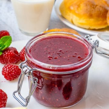 A glass jar of glossy Raspberry Curd surrounded by fresh raspberries with brioche buns and milk behind it.