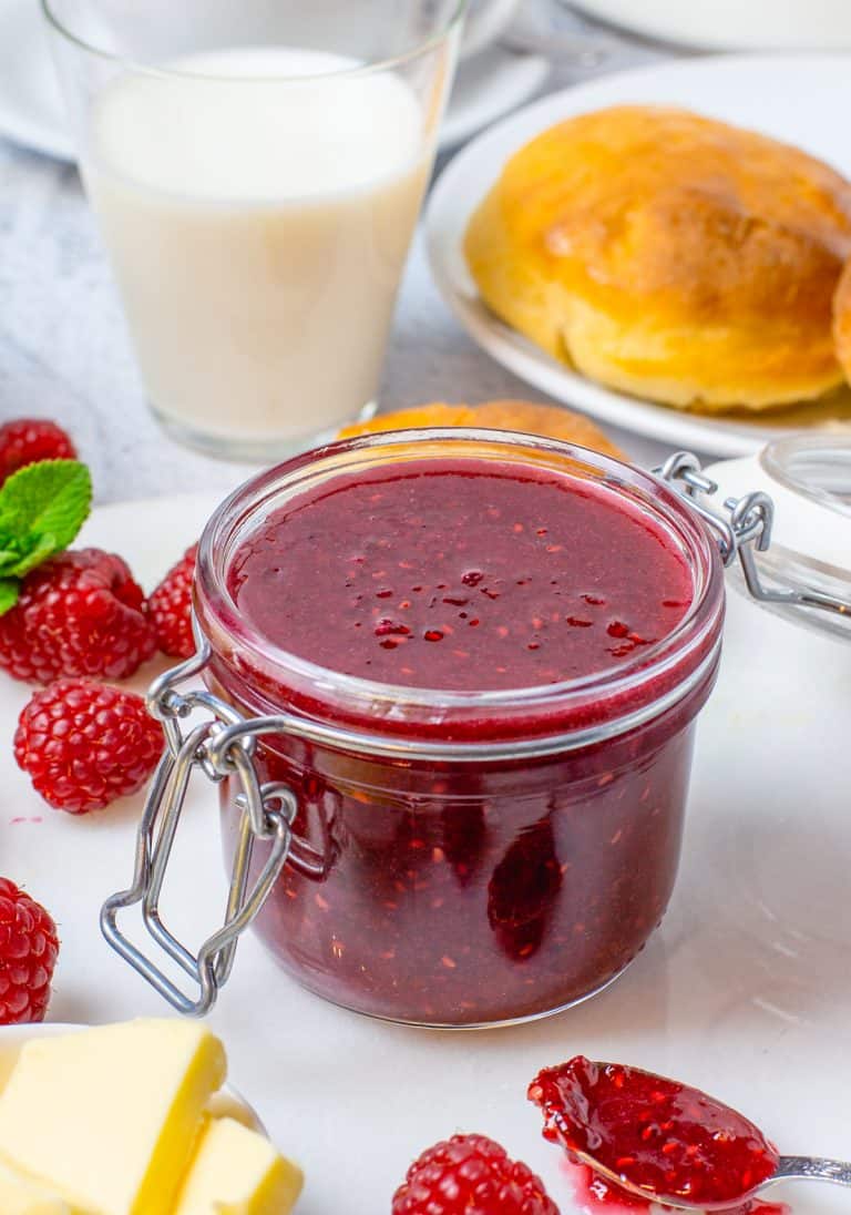A glass jar of glossy Raspberry Curd surrounded by fresh raspberries with brioche buns and milk behind it.
