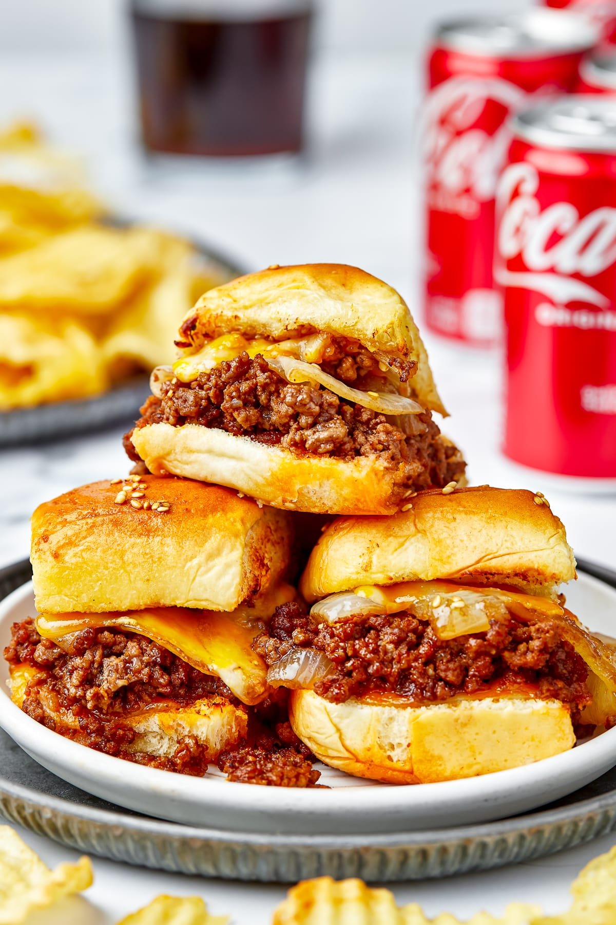 Sloppy Joe Sliders stacked on a white plate that is sitting on a metal serving platter, wavy chips and cans of coca-cola in the background