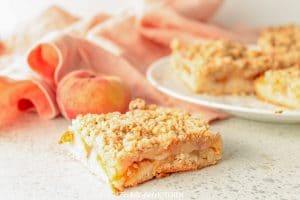 One Peach Crumble Bar in font of peach and plate of bars