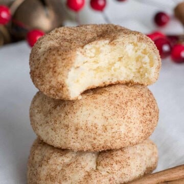 Three stacked cinnamon sugar Cream Cheese Cookies with a bite taken out of the top cookie, with festive red berries and a cinnamon stick in the background.