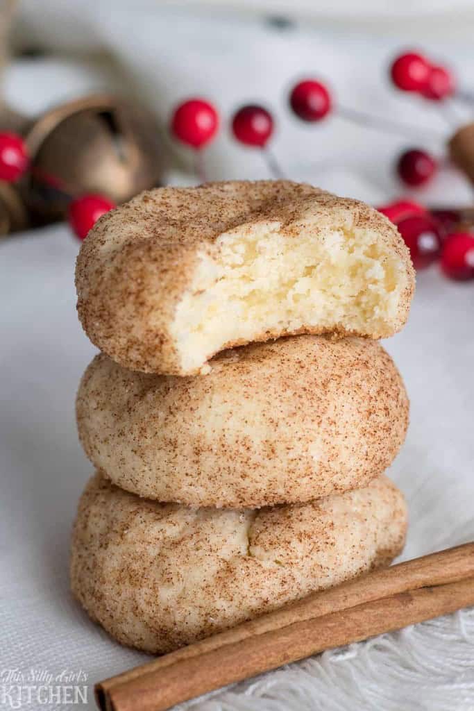 Three stacked cinnamon sugar Cream Cheese Cookies with a bite taken out of the top cookie, with festive red berries and a cinnamon stick in the background.