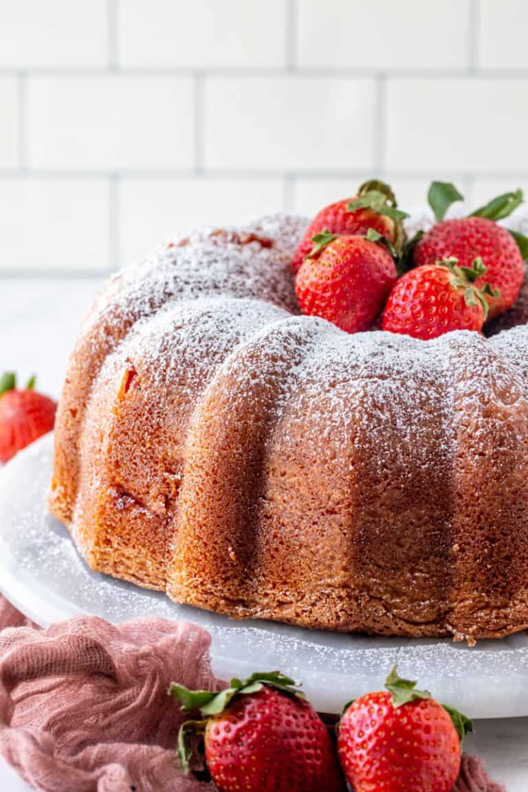 Easy Strawberry Cheesecake Bundt Cake Recipe finished on a white cake stand with powdered sugar dusting and fresh strawberries arranged on a pink linen.