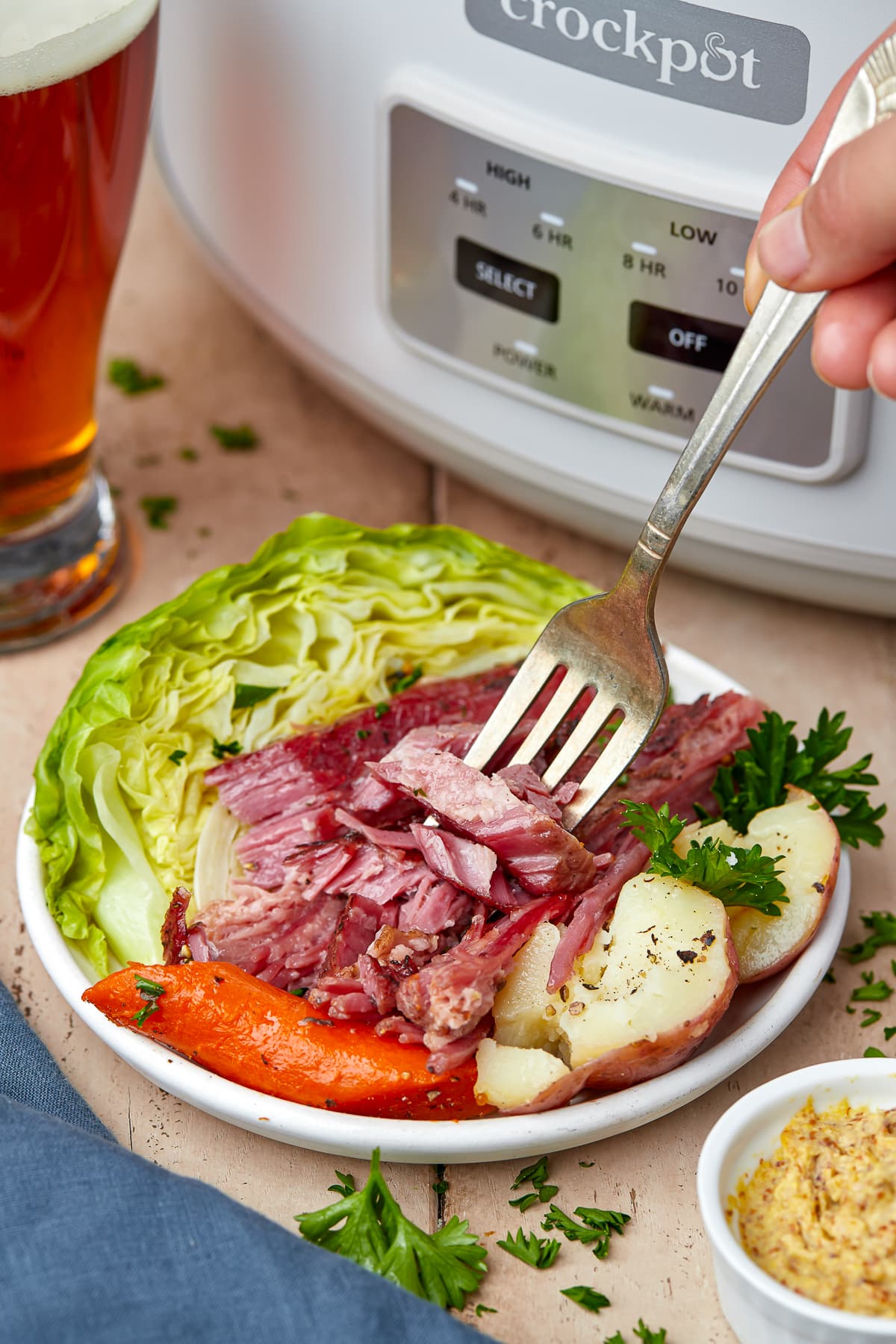 Plated shot of slow cooker corned beef and cabbage on a white plate with white slow cooker in the background, glass of beer, on a wooden backdrop