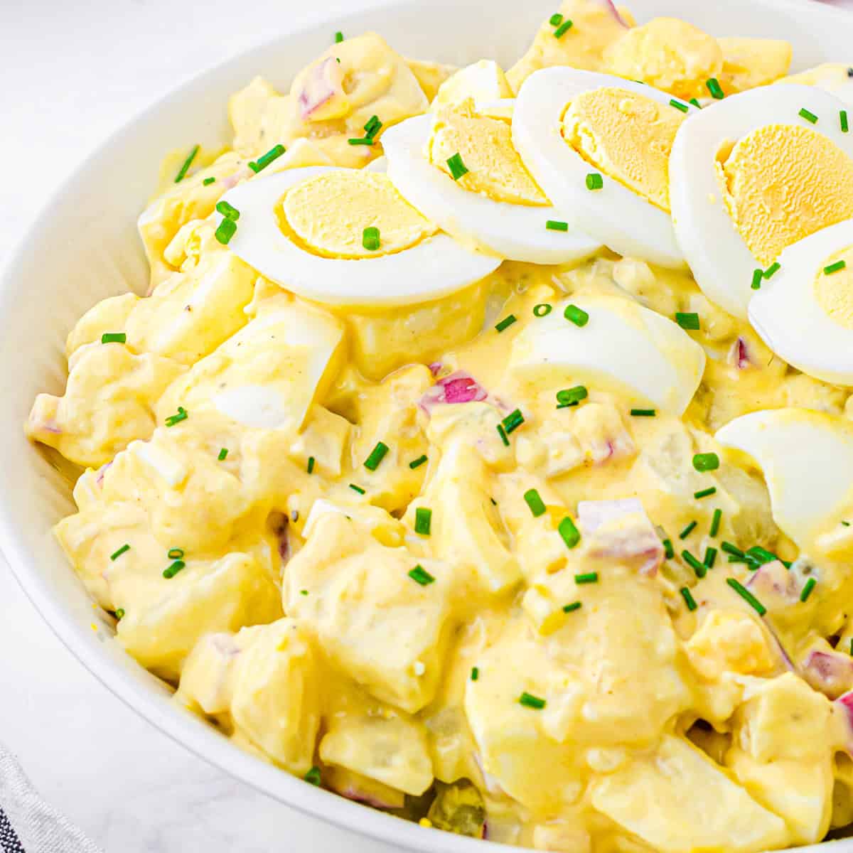 Square close-up of creamy classic potato salad with hard-boiled eggs and fresh chives in a white bowl.