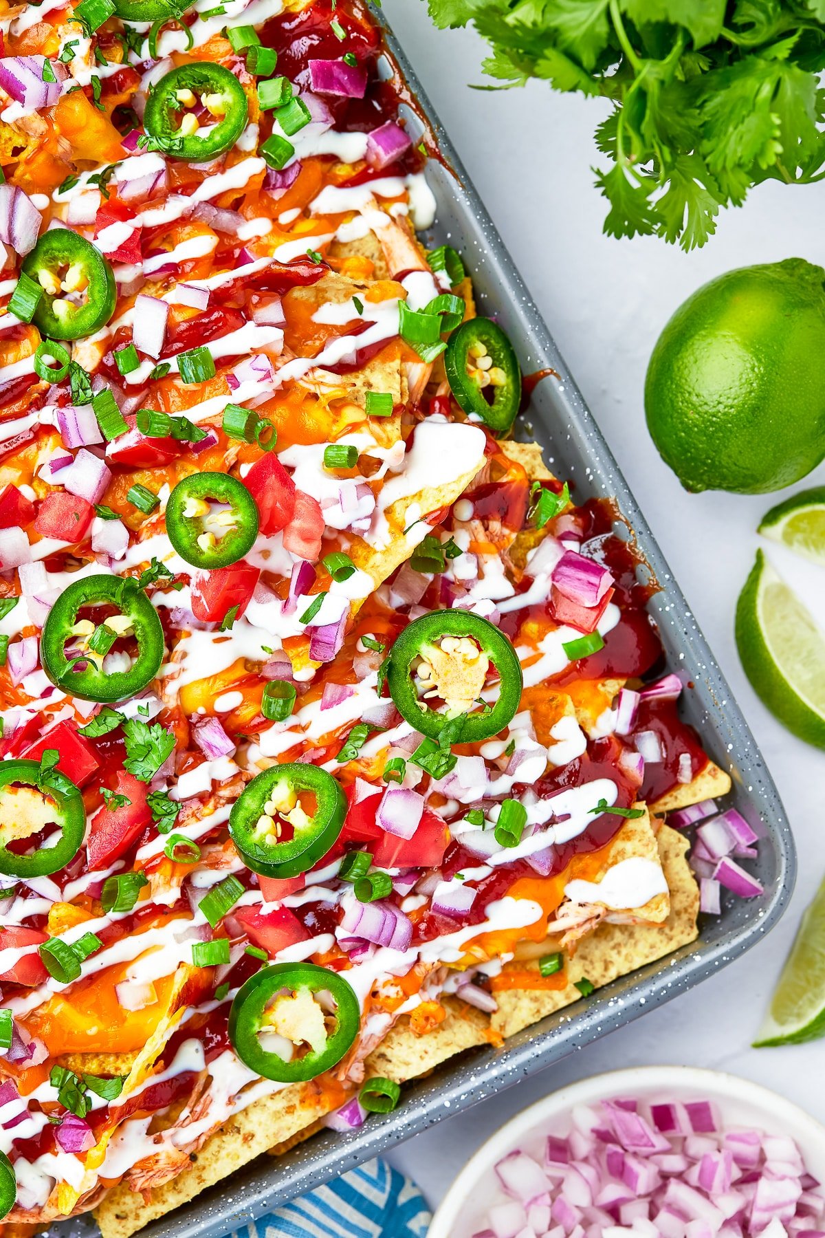 Upclose overhead shot of a sheet tray covered with BBQ Chicken Nachos on a white stucco tabletop with limes and red onion