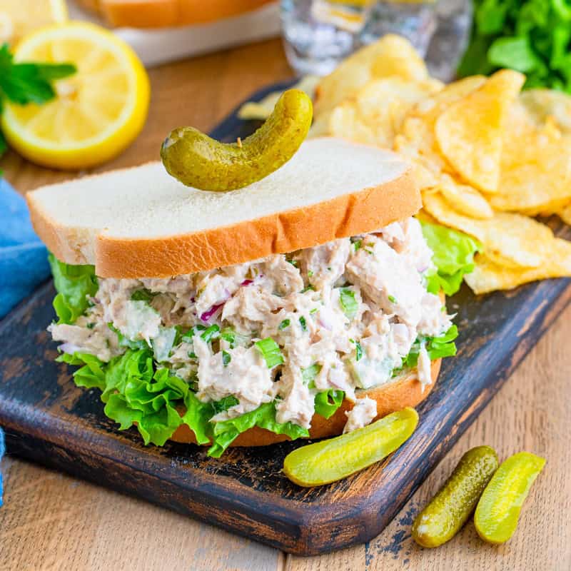 A tuna salad sandwich on white bread with lettuce, served with pickles and chips.