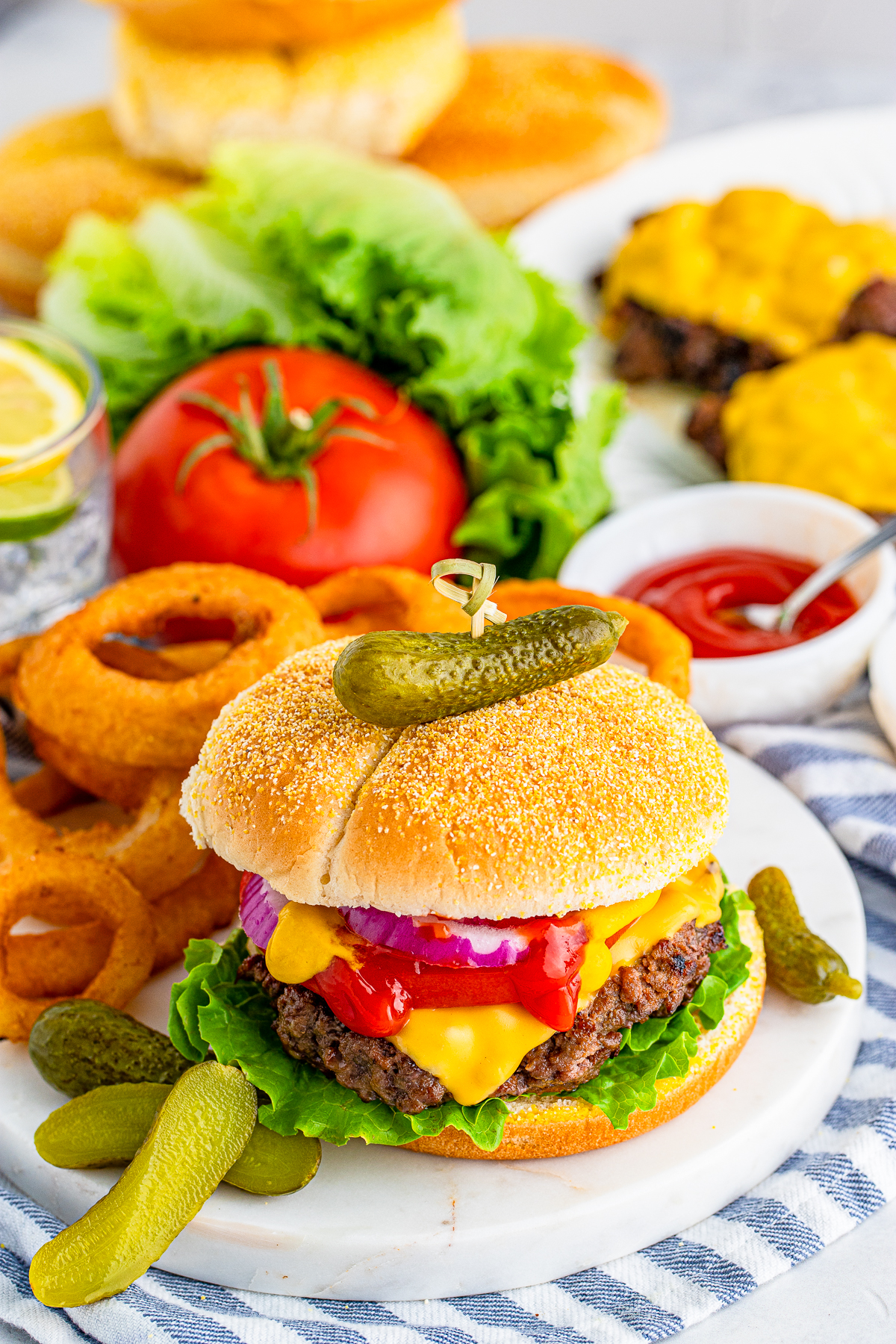 Cheeseburger on marble board with onion rings, pickles and more burgers and buns in background
