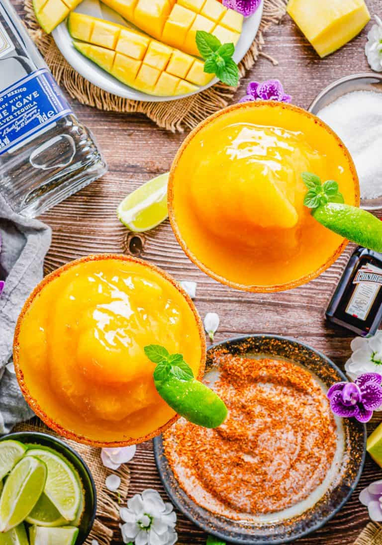 A frozen mango margarita with a chili lime salt rim and a lime wedge on a wooden board.