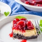 A cherry cheesecake bar on a white plate with glossy cherry pie filling spilling over the creamy layer and Oreo crust.