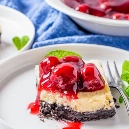 A cherry cheesecake bar on a white plate with glossy cherry pie filling spilling over the creamy layer and Oreo crust.