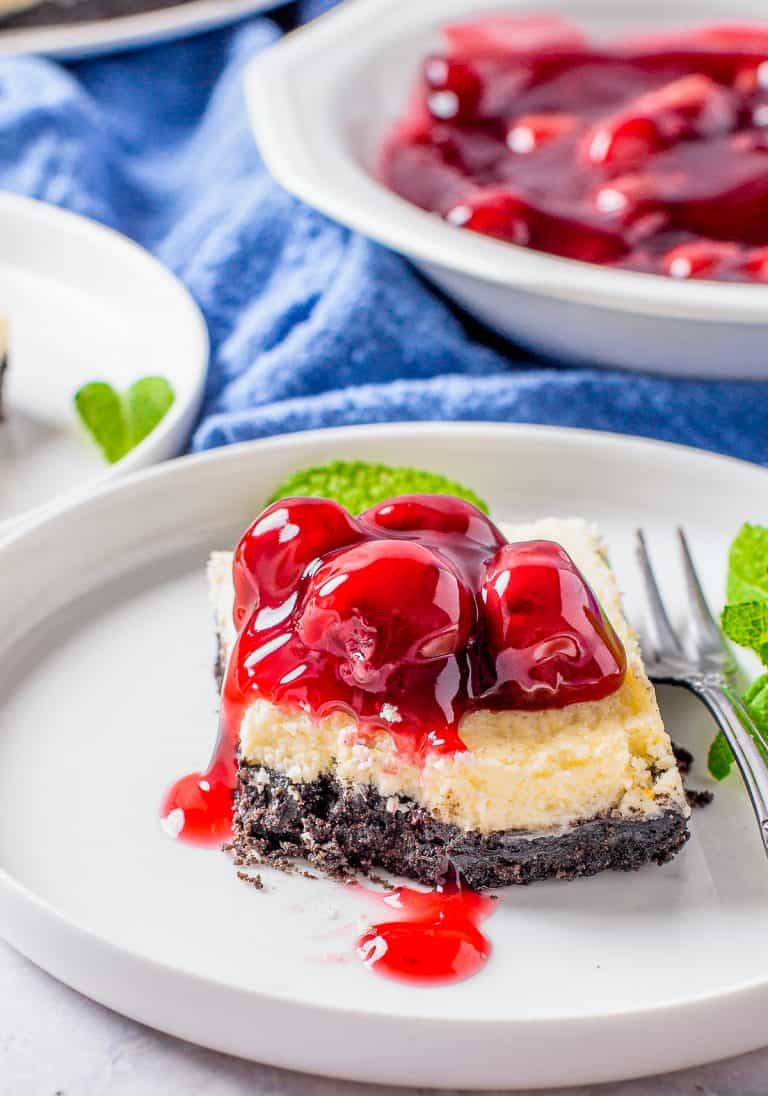 A cherry cheesecake bar on a white plate with glossy cherry pie filling spilling over the creamy layer and Oreo crust.
