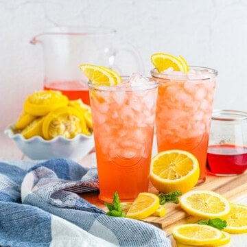Two glasses of homemade pink lemonade over ice with lemon slices and a pitcher in the background.