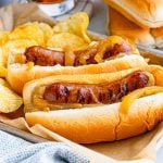 Square image of two brats in buns on tray with chips.
