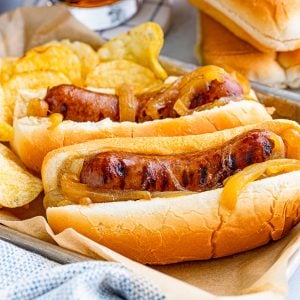 Square image of two brats in buns on tray with chips.