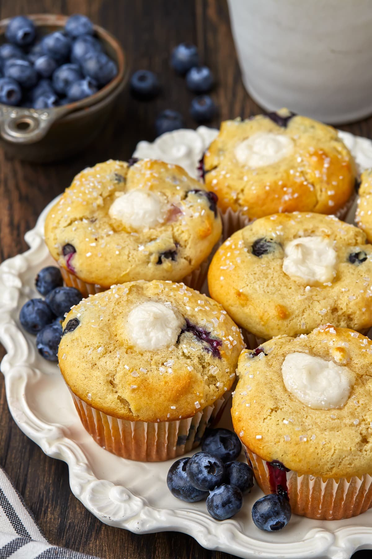 Blueberry cream cheese muffins sitting on a decorative white plate on a dark wooden tabletop with a measuring cup full of blueberries