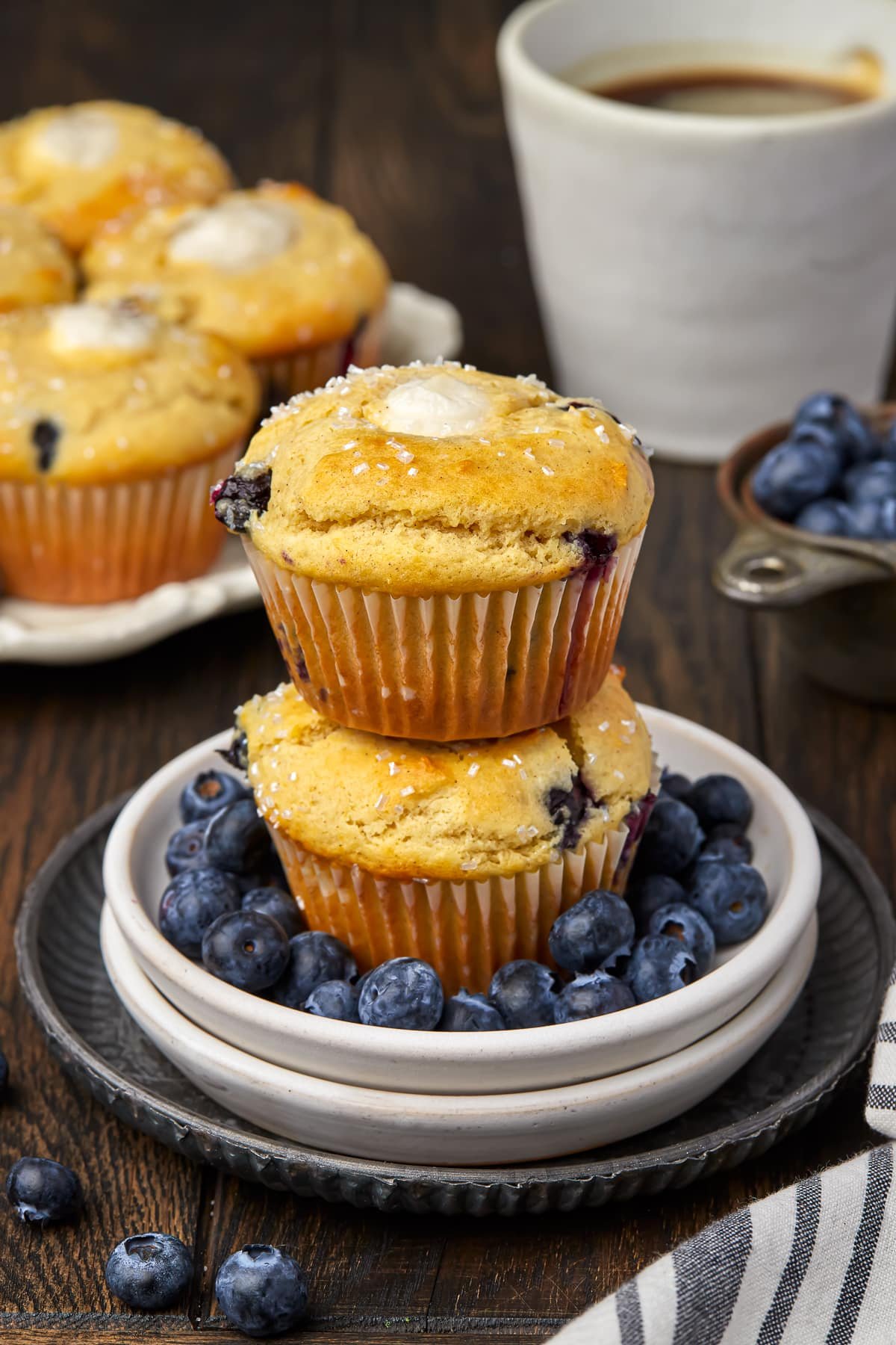 2 blueberry cream cheese muffins stacked on top of each other on small white plates and a metal serving plate with bluerries scattered around. A plate of muffins in the background and cup of black coffee.