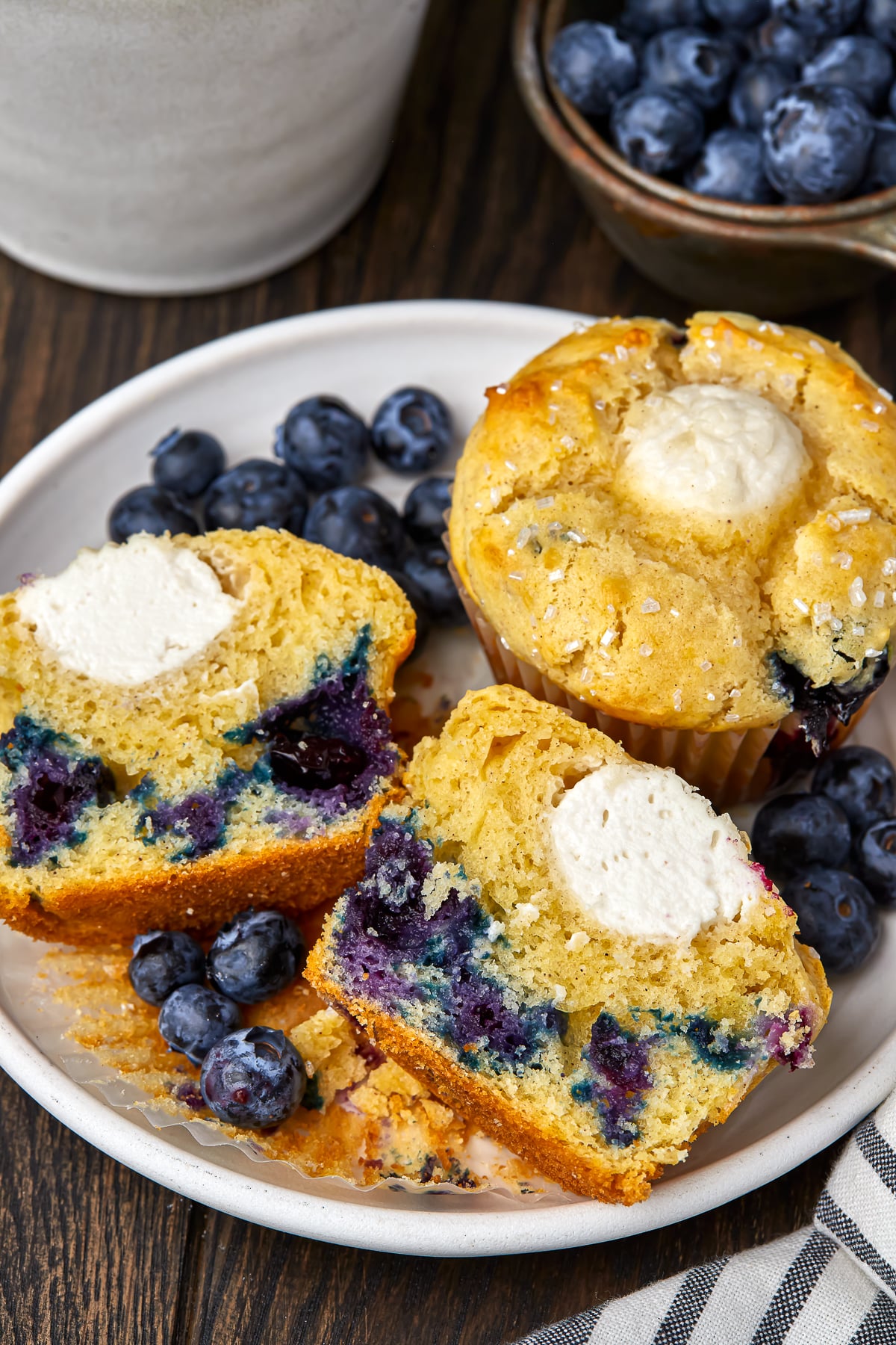 A blueberry cream cheese muffin cut in half to show the filling on a white plate with blueberries on a dark wooden tabletop