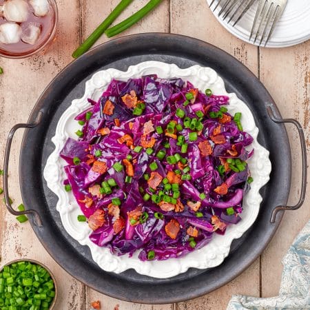 A black bowl of sweet and sour cabbage topped with crispy bacon and sliced scallions.