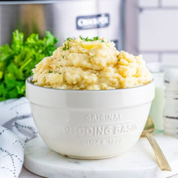 A white bowl of creamy slow cooker mashed potatoes topped with melting butter and fresh parsley.