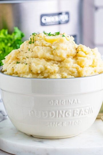 A white bowl of creamy slow cooker mashed potatoes topped with melting butter and fresh parsley.