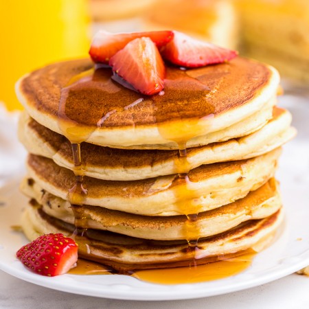 Square image of stacked pancakes on plate with syrup dripping down side.
