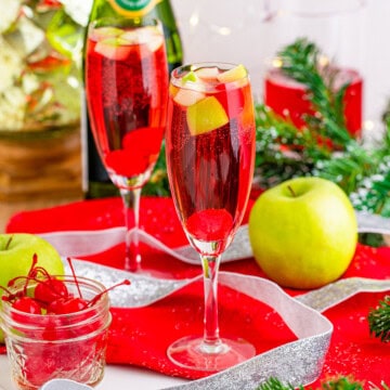 Two champagne flutes of sparkling red Christmas Mocktails garnished with cherries and green apple.