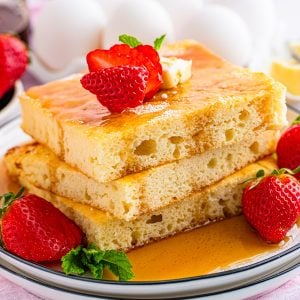 Square image of stacked pancakes with syrup, butter and strawberries.