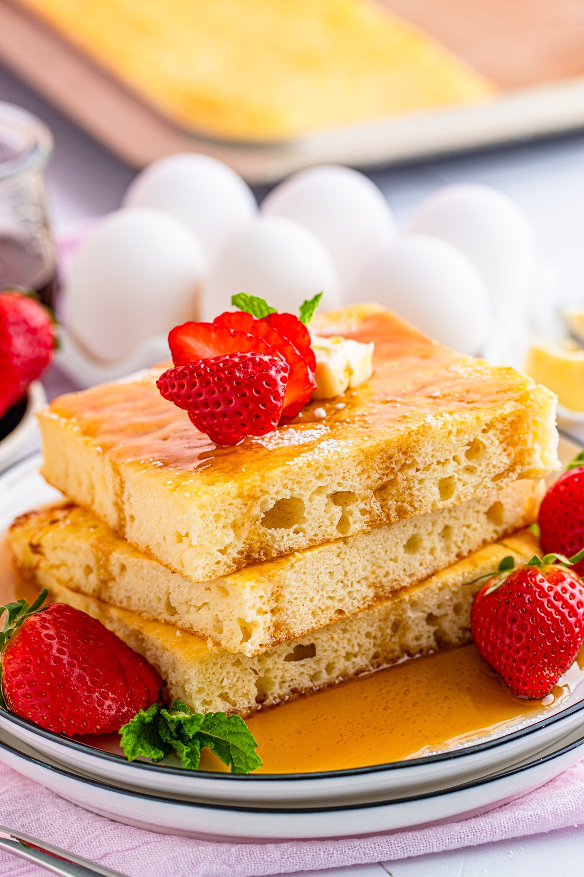 Three stacked Sheet Pan Pancakes sliced on plate with syrup and strawberries.