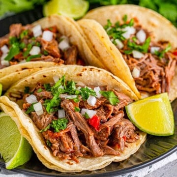 A plate of slow cooker shredded beef tacos topped with cilantro, onion, and lime.