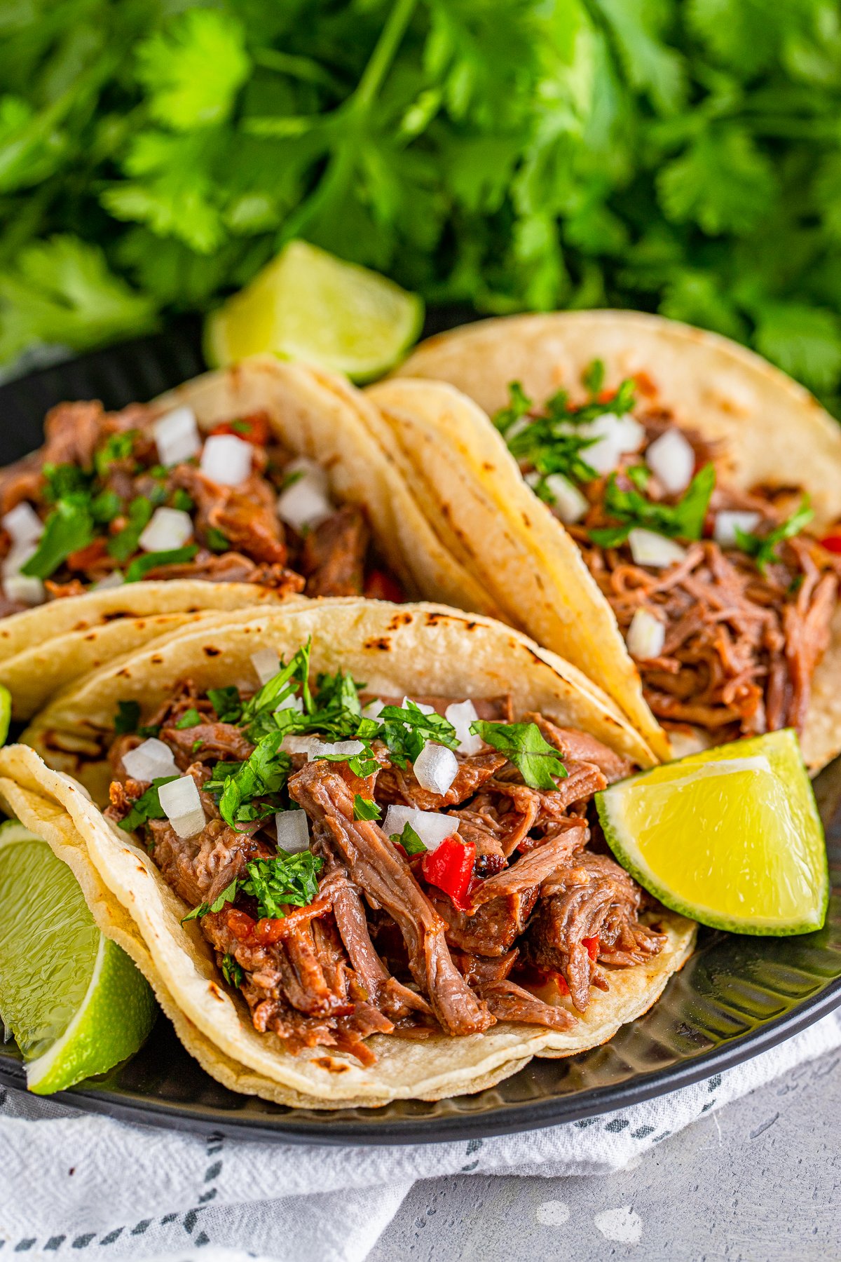 Three Shredded Beef Tacos on black plate with toppings and lime garnishes.