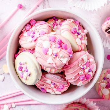 A bowl of chocolate covered Oreos coated in pink and white chocolate with colorful sprinkles.