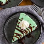 A slice of no bake mint chocolate cheesecake on a plate with whipped cream and chocolate drizzle.