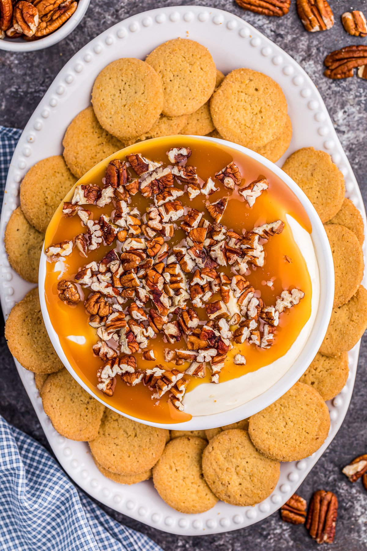 Overhead photo of Butterscotch Cheesecake Dip in bowl with sauce and chopped nuts with cookies surrounding it.