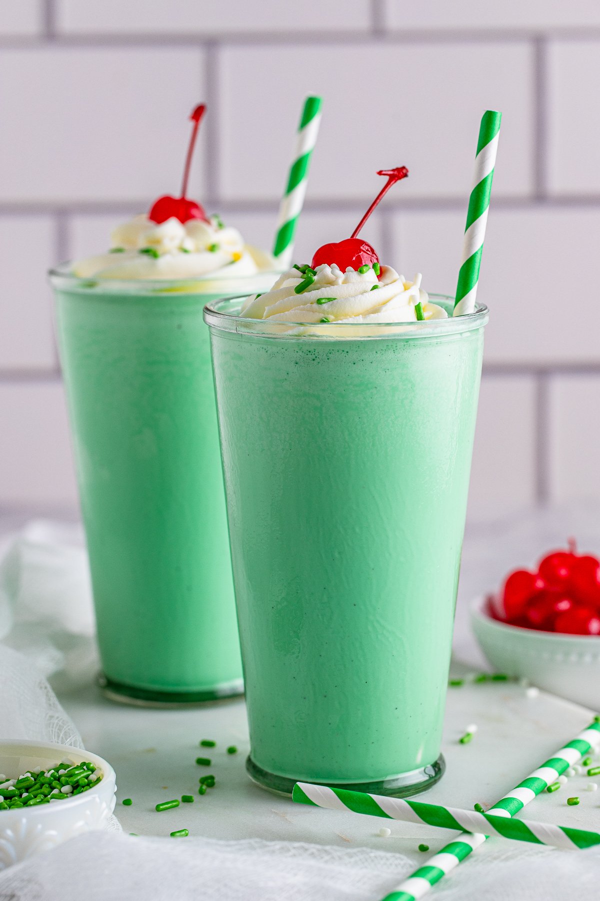 Photo of two Shamrock Shakes in clear glasses side by side with green and white swirl straws served with whipped cream and a cherry on top - by This Silly Girl's Kitchen.